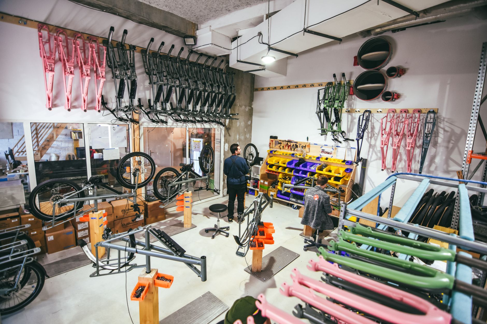 atelier de vélos à Bordeaux avec des cadres colorés accrochés au mur, des pieds d’atelier et deux personnes au travail